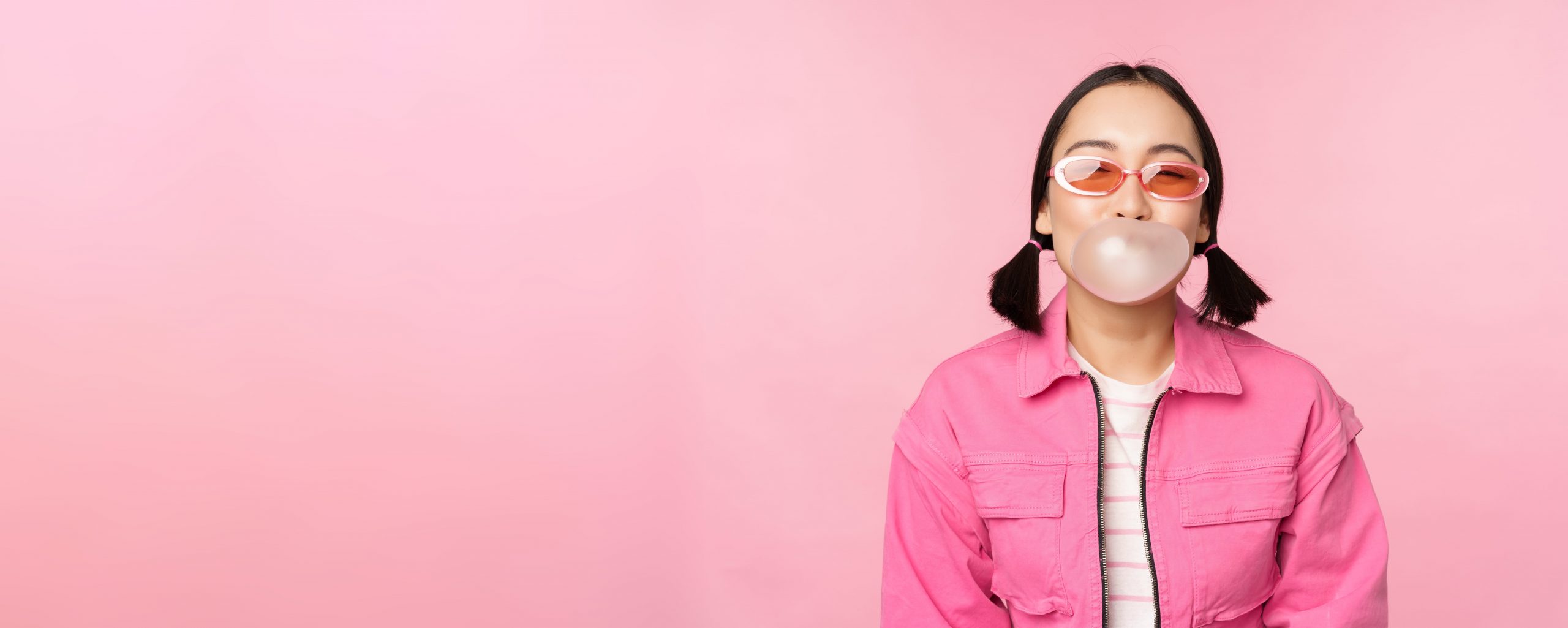 A girl blowing bubble gum against a pink background.